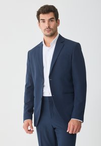 Σκούρο μπλε σακάκι κοστουμιού, μονόπετο με δύο κουμπιά, πέτο με σχισμή και τσέπη στο στήθος. Συνδυάζεται με λευκό πουκάμισο με κουμπιά.