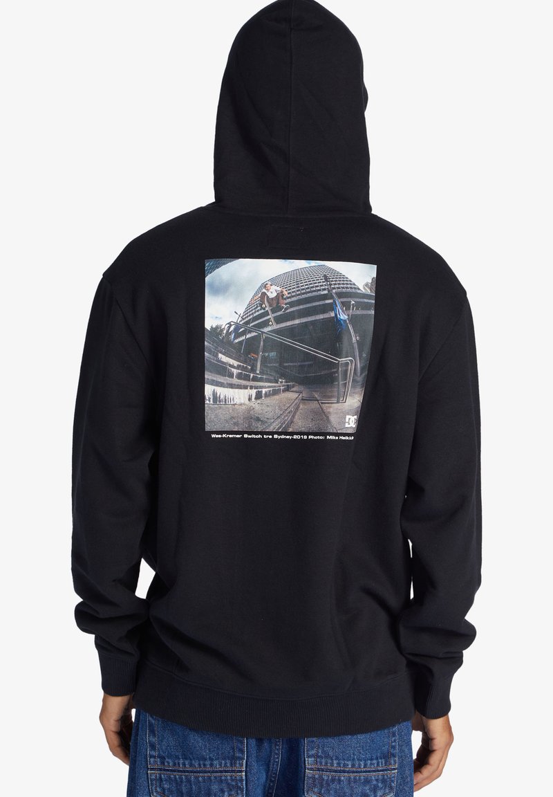 Sort hættejakke med et stort grafisk print på ryggen, som viser en skateboarder. Blødt stof, standard pasform, med ribbede manchetter og kant.