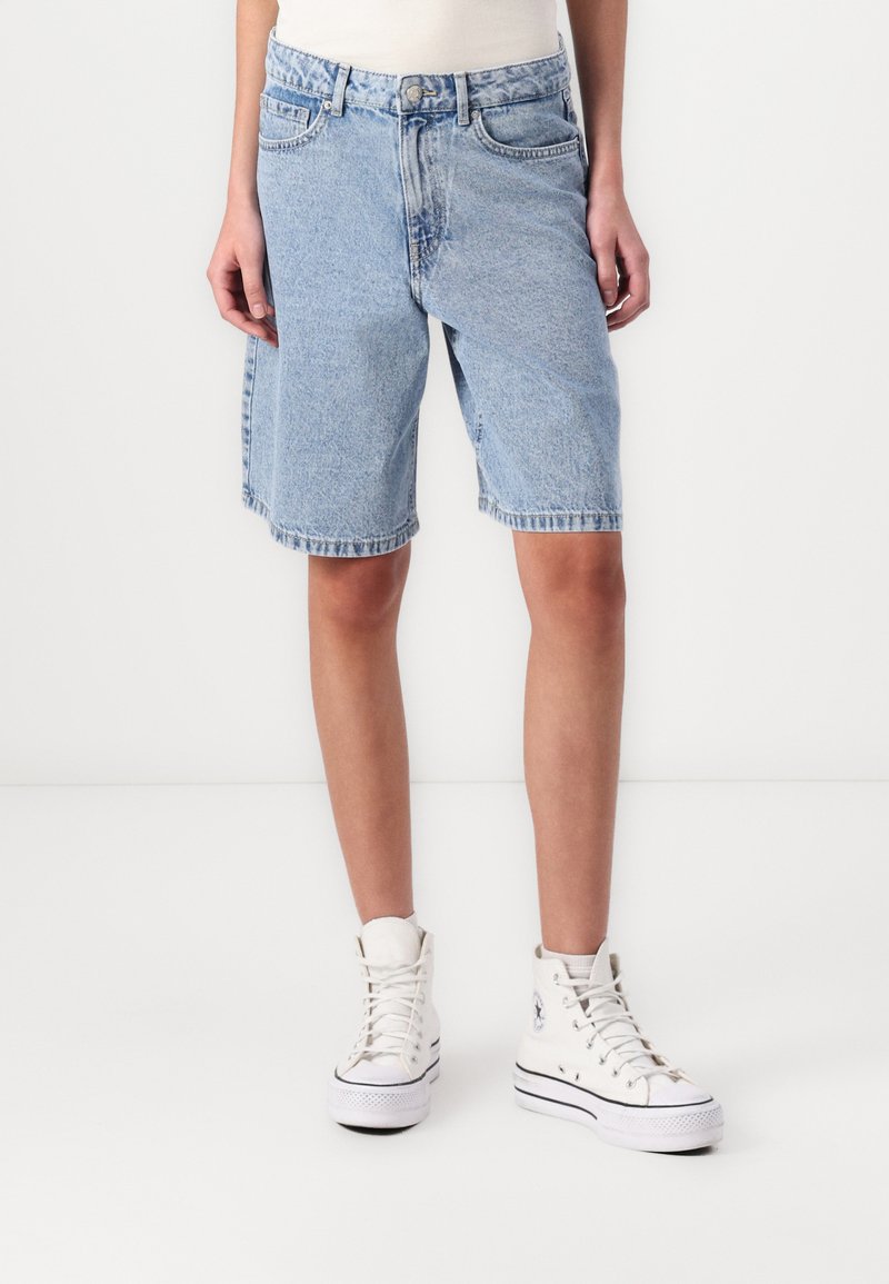 Light blue denim shorts with a loose fit, cuffed hem, five pockets, and a button closure. Paired with white high-top sneakers.