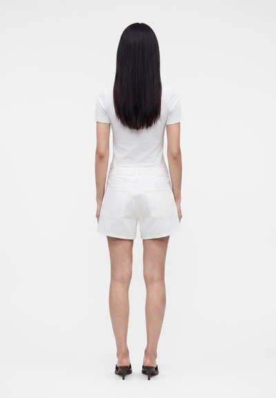 Γυναίκα με μακριά μαύρα μαλλιά, φορώντας λευκό κοντομάνικο μπλουζάκι, λευκά σορτς και μαύρα σανδάλια με τακούνι, στέκεται γυρισμένη με την πλάτη σε λευκό φόντο.