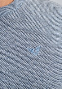 Pull tricoté bleu clair texturé avec un col rond et un petit emblème brodé représentant des ailes en bas à gauche.