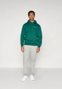 Zöld kapucnis pulóver húzózsinórokkal és elülső zsebbel, világosszürke melegítőnadrággal és fehér sportcipővel párosítva; laza szabás és hétköznapi stílus.