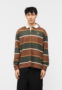 Νέος άνδρας που φοράει πουλόβερ πολο με μακριά μανίκια σε καφέ, πράσινες και πορτοκαλί ρίγες, με λευκό γιακά και μαύρο παντελόνι, στέκεται με το πρόσωπο προς τα εμπρός.