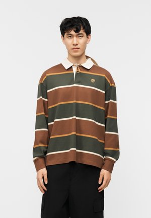 Ung mann iført en langermet polotrøye med brune, grønne og oransje striper, hvit krage og svarte bukser, står vendt mot kameraet.