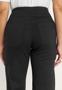 Nainen käyttää korkeavyötäröisiä mustia housuja, joissa on takataskut, ja seisoo rentoina käsivarret sivuilla yksinkertaista taustaa vasten.