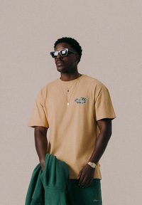 T-shirt en coton beige avec un logo circulaire à motifs camouflage. Associé à un pantalon de survêtement vert et un sweatshirt à capuche assorti, agrémenté de lunettes de soleil.