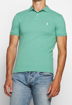 Mann trägt ein tailliertes, mintgrünes Poloshirt mit kurzen Ärmeln und hellblaue Jeans, steht vor einem einfarbigen Hintergrund.