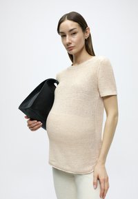 Haut tricoté rose clair à manches courtes, col rond, finition texturée, associé à un sac fourre-tout en cuir noir tenu dans une main.