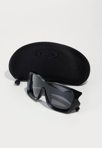 Black sports sunglasses with a sleek, angular design and grey lenses, placed next to a solid black protective case featuring an embossed logo.