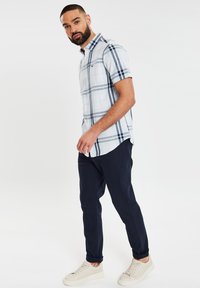 Kurzärmliges, knöpfbares Hemd im hellblauen Karo-Muster mit dunkelblauen und weißen Akzenten, kombiniert mit dunkelblauen, schmal geschnittenen Hosen und beigen Schuhen.