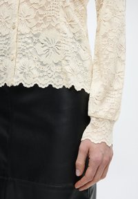 Bluză cu mâneci lungi din dantelă florală crem, cu margini ondulate, asortată cu o fustă din piele neagră, mâna relaxată lângă corp.