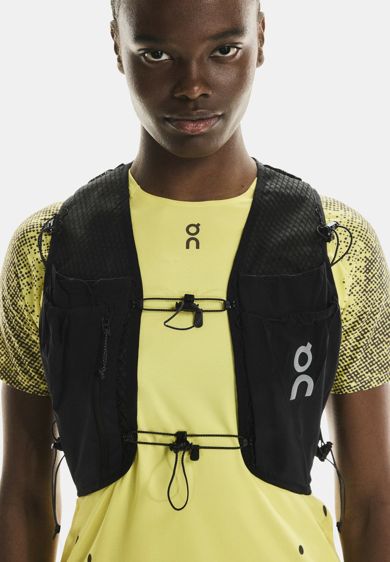 Gros plan d'une personne portant un t-shirt sportif jaune et un gilet de course noir avec des poches avant et des cordons réglables.