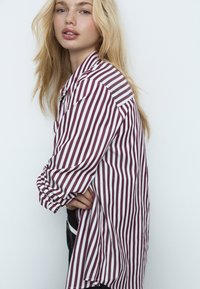 Chemise boutonnée à rayures bordeaux et blanches, présentant une coupe décontractée, des manches longues et une poche poitrine, portée avec un jean.