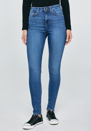 Modré džíny se štíhlým střihem nošené s černým dlouhým rukávem a černými nízkými teniskami na jednobarevném světlém pozadí.