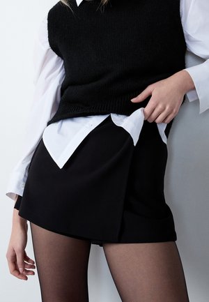 Person iført sort ærmeløs strikvest over hvid skjorte med knapper, sort nederdel og gennemsigtige sorte strømpebukser, læner sig op ad en lys grå væg.