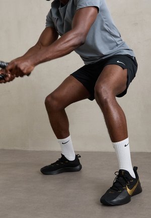 Mann in grauem Shirt und schwarzen Nike-Shorts und Schuhen macht eine Kniebeuge und hält ein Seil in einem Indoor-Trainingsbereich.