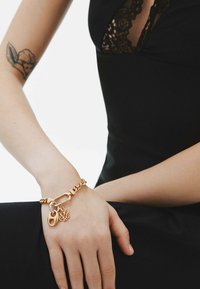 Pulsera en tono dorado con una cadena gruesa, que presenta un candado y un charm floral. Textura suave con acentos brillantes, llevada en una muñeca contra un atuendo negro.