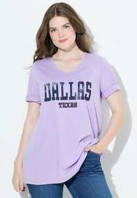 Mujer joven con cabello largo castaño, vistiendo una camiseta light púrpura de "Dallas Texas" y jeans azules, posando con una mano en el bolsillo.