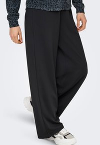 Άτομο που φοράει φαρδιά μαύρη παντελόνα, λευκά αθλητικά παπούτσια και μπλε πουκάμισο με λεοπάρ σχέδιο και μακριά μανίκια, στέκεται με το ένα πόδι ελαφρώς λυγισμένο.