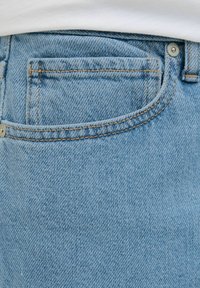 Gros plan d'une poche de jean en denim bleu clair avec coutures visibles et rivet métallique sur un fond de tissu blanc.