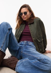 Donna seduta sul pavimento che indossa una giacca verde scuro, una maglia bordeaux, jeans blu, stivali marroni e occhiali da sole aviator su uno sfondo bianco e semplice.