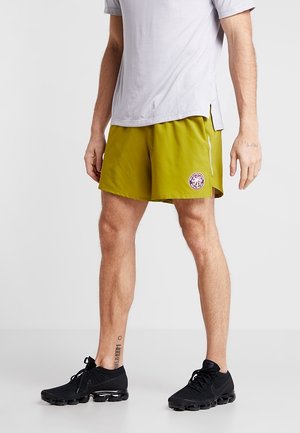 Mand iført olivengrønne sportsshorts med et cirkulært logo, lysegrå t-shirt og sorte løbesko, stående på en hvid baggrund.