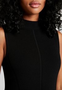 Gros plan sur une personne avec des cheveux noirs bouclés portant un haut en tricot sans manches de couleur noire avec un col montant.