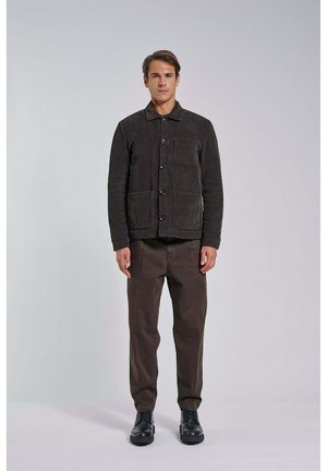 Homme debout portant une veste en velours côtelé marron foncé à boutons, un pantalon marron assorti et des chaussures en cuir noir sur un fond clair uni.