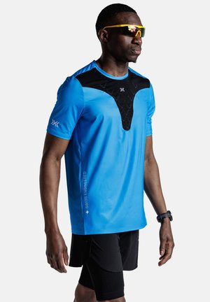 Atleet die een blauw-zwart sportshirt, zwarte short, gele reflecterende zonnebril en een zwart polshorloge draagt, staand tegen een witte achtergrond.