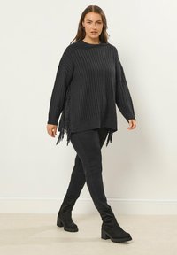 Kobieta spacerująca w pomieszczeniu, ubrana w luźny czarny sweter z prążkowanym wzorem i frędzlami po bokach, czarne skinny pants oraz czarne botki na obcasie.