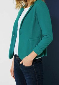 Teal blazer i texturerat tyg; har två fickor, en enkel knappstängning och uppvikt ärm. Bärs över en vit topp och mörka jeans.
