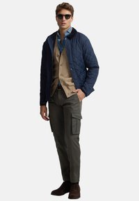 Chaqueta acolchada azul marino con cuello negro, cárdigan marrón claro, camisa azul, pantalones cargo oliva y botas marrones. Los accesorios incluyen gafas de sol.
