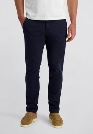 Mann trägt dunkelblaue, eng anliegende Hose, weißes Hemd und hellbraune Wildleder-Loafer, steht mit entspannten Händen.