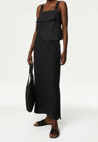 Tenue noire en deux pièces composée d'un haut sans manches à couches avec de larges bretelles et d'une longue jupe ajustée. Le mannequin porte un sac tissé noir.