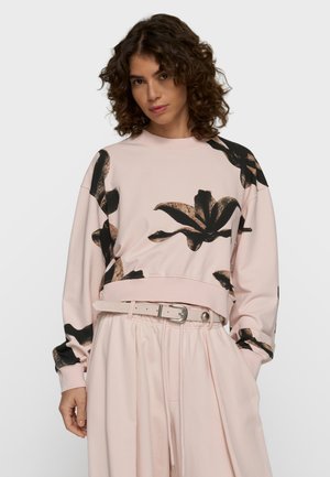 Femme aux cheveux bouclés portant un sweat-shirt rose clair avec de grands motifs floraux noirs et bruns et un pantalon taille haute assorti avec une ceinture.