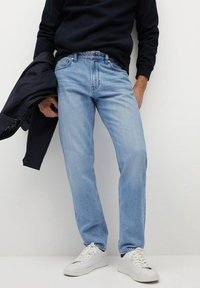 Jean en denim bleu clair à coupe droite, avec un design standard à cinq poches et une légère décoloration. Porté avec un pull bleu marine et des baskets blanches.