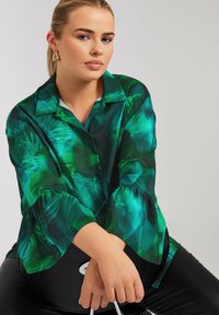 Blusa verde con un patrón de hojas, mangas largas con puños acampanados y cierre de botones en el frente. Combinada con pantalones negros, descansando sobre una superficie.