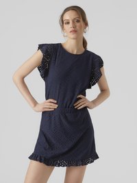 Naine, kes kannab mereväesinist tikk-kangast pitskleiti volangiliste varrukate ja äärega, seisab käed puusadel halli tausta ees.