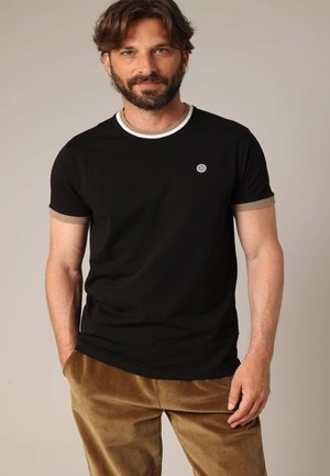 Zwarte t-shirt met korte mouwen en een witte kraag, met tan accenten op de mouwen, voorzien van een klein geborduurd logo op de borst. Corduroy broek.