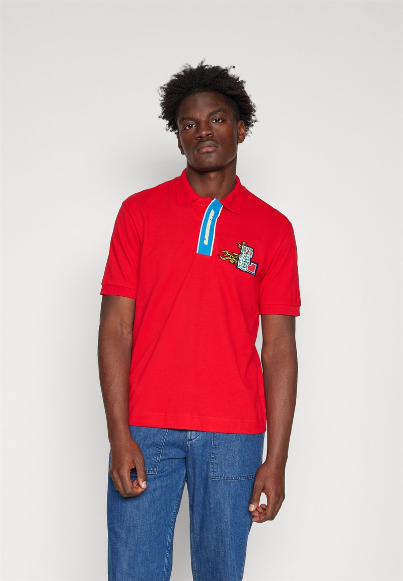 T-shirt polo rouge avec un col bleu, présentant un design brodé sur la poitrine avec des flammes et des accents à motif damier.