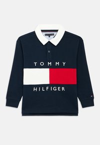 Temno modra polo majica z dolgimi rokavi in belim ovratnikom. Ima vodoravno pasico z rdečimi in belimi odseki, na kateri je napis "TOMMY HILFIGER".