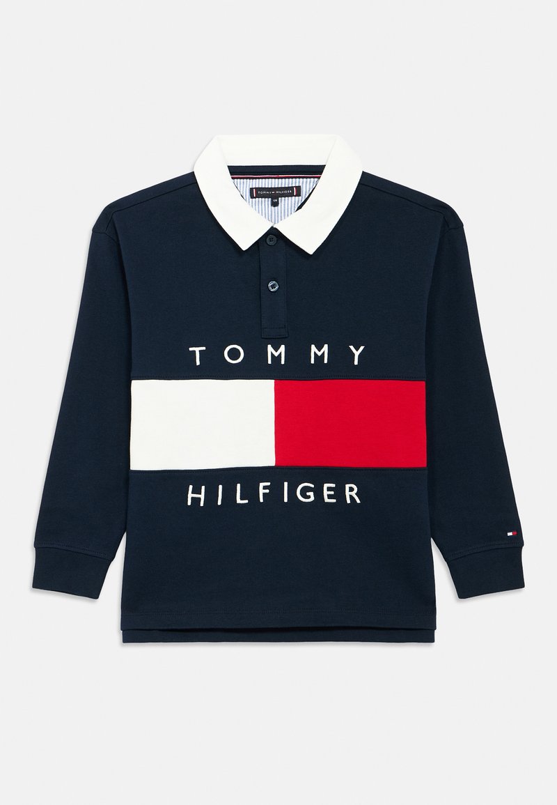Temno modra polo majica z dolgimi rokavi in belim ovratnikom. Ima vodoravno pasico z rdečimi in belimi odseki, na kateri je napis "TOMMY HILFIGER".