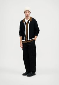 Fekete kardigán fehér és barna díszítéssel, gombosan lecsukva, barna galléros ing fölött viselve. Fekete cargo nadrággal és fekete cipővel párosítva.