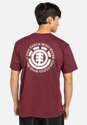 Camiseta de algodón en color marrón con un gráfico circular blanco que presenta palabras elementales y un logo en la parte de atrás. Diseño de mangas cortas y cuello redondo.