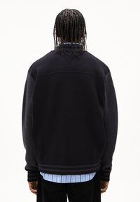 Personne avec des cheveux tressés portant un pull noir et gris foncé par-dessus une chemise à rayures bleues, de dos contre un fond blanc uni.