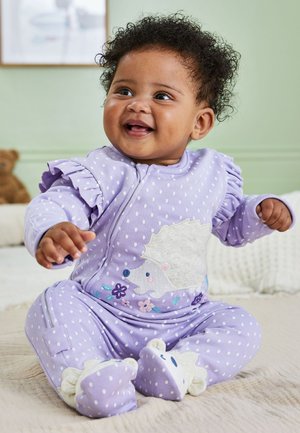 Bambino sorridente seduto su un letto che indossa una tutina lavanda a pois con disegni di ricci e fiori, che porge entrambe le mani in avanti.