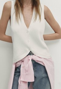 Cardigan bianco a righe senza maniche con scollatura a V profonda e tre bottoni, abbinato a una camicia rosa annodata e jeans azzurri a vita alta.