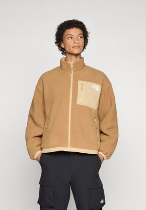 Brun fleecejakke med høy krage, glidelås lomme på brystet, og elastiske mansjetter, med kontrasterende trim og logo på lommen.