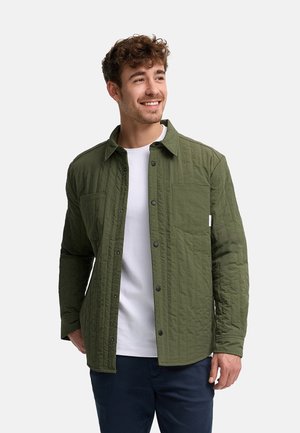 Hombre sonriente con camisa verde acolchada de botones sobre camiseta blanca y pantalones oscuros, de pie contra un fondo claro y liso.