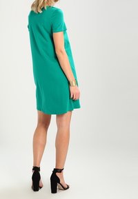 Vrouw in een groen jurkje met korte mouwen, zwarte enkellaarsjes met een enkelbandje en een gouden manchette, staande in zijaanzicht op een effen achtergrond.